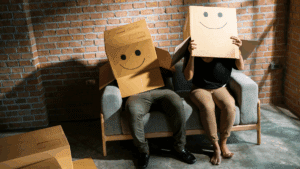 Un couple assis sur n canapé, leur tête dans des boites en carton avec des sourires dessinés dessus.