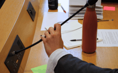 Une main saisie un micro pour intervenir sur un bureau officiel encombré de papiers de notes et d'une gourde.