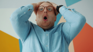 Un homme en chemise bleu avec des lunettes se prend la tête dans les mains surpris pris de face.