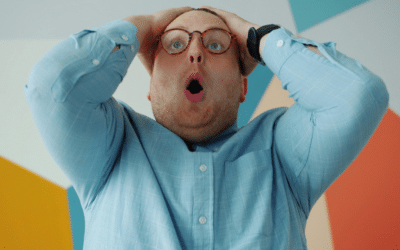 Un homme en chemise bleu avec des lunettes se prend la tête dans les mains surpris pris de face.